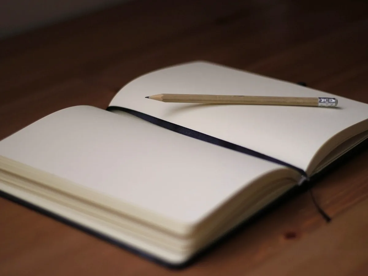 An open notebook with blank cream pages and a dark ribbon marker resting on a warm wooden desk, a pencil lying across the page