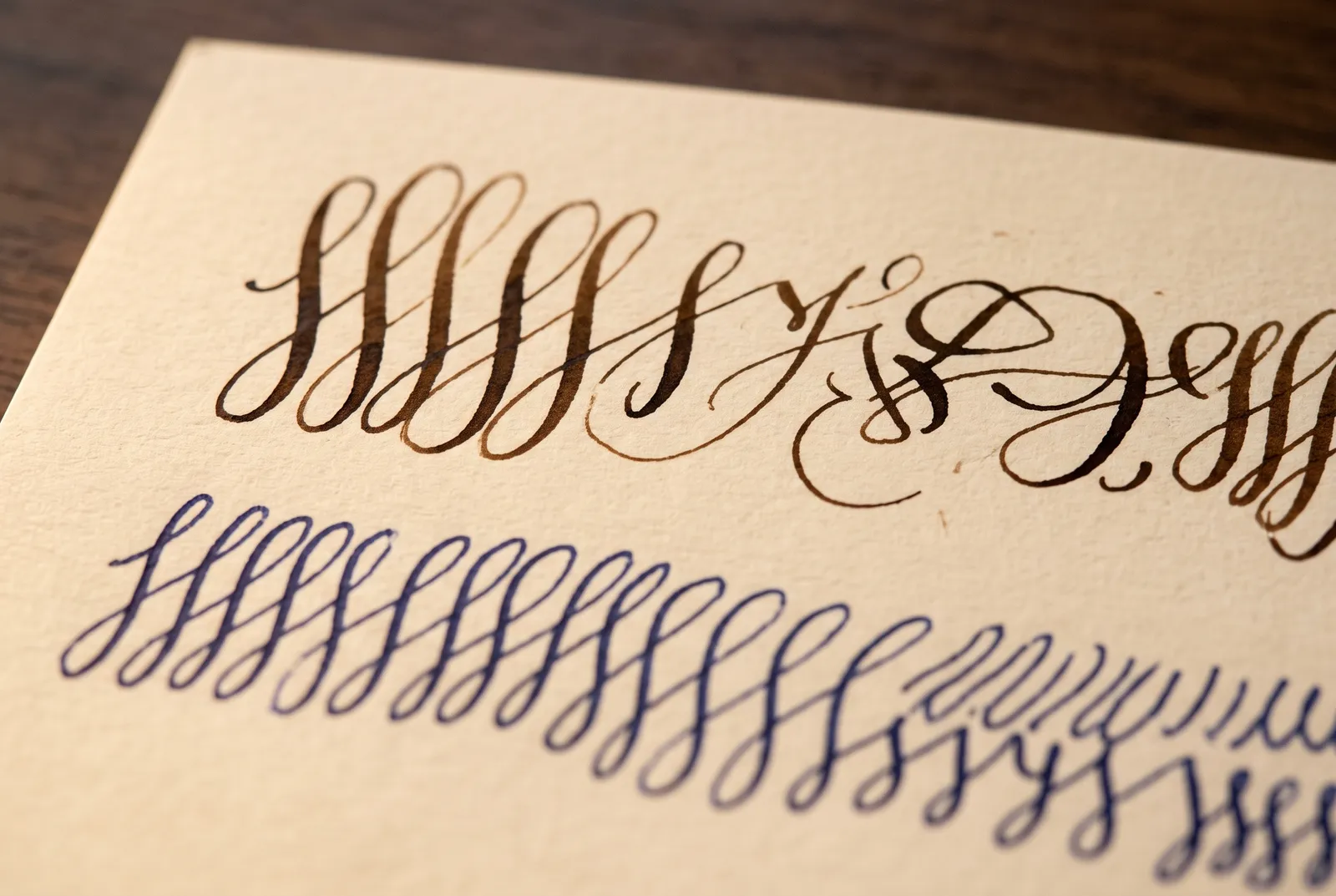 Two rows of practice pen strokes on cream paper: looping fountain-pen flourishes above and uniform ballpoint loops below