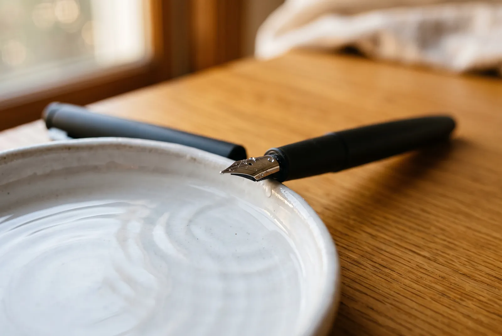 A fountain pen resting nib-down against the rim of a white ceramic dish of water, droplets on the steel nib